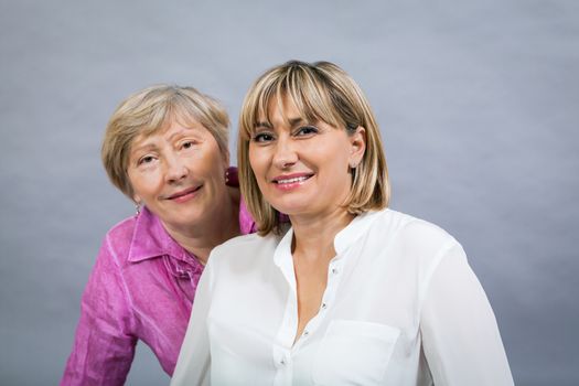 Attractive stylish blond senior lady with her beautiful middle-aged daughter posing together with her hands on her shoulders smiling at the camera on a grey studio background