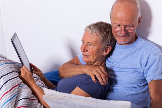Senior Couple Relaxing in Bed with Tablet Computer and Newspaper