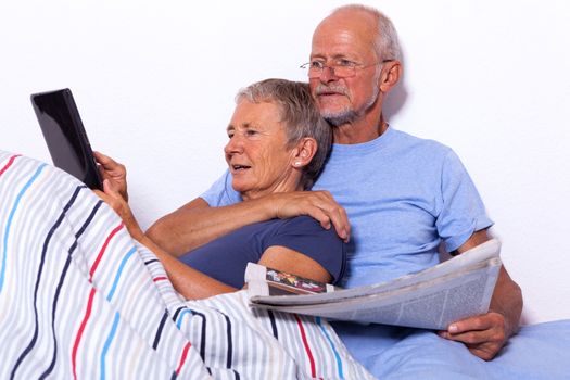 Senior Couple Relaxing in Bed with Tablet Computer and Newspaper