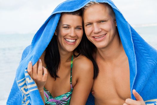 Cheerful Caucasian young happy couple made of a blond handsome man and an attractive brunette woman, with a blue towel covering their heads
