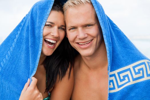 Cheerful Caucasian young happy couple made of a blond handsome man and an attractive brunette woman, with a blue towel covering their heads