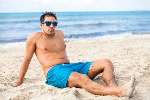 Handsome man wearing trendy sunglasses and his swimsuit relaxing on the beach sitting on the golden beach sand soaking up the summer sun with the ocean behind him