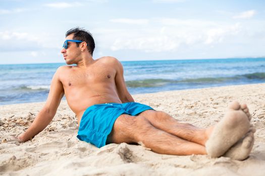Handsome man wearing trendy sunglasses and his swimsuit relaxing on the beach sitting on the golden beach sand soaking up the summer sun with the ocean behind him