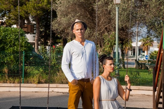 Elegant trendy young couple in fashionable modern clothes and accessories posing outdoors in the summer sunshine in a garden or park