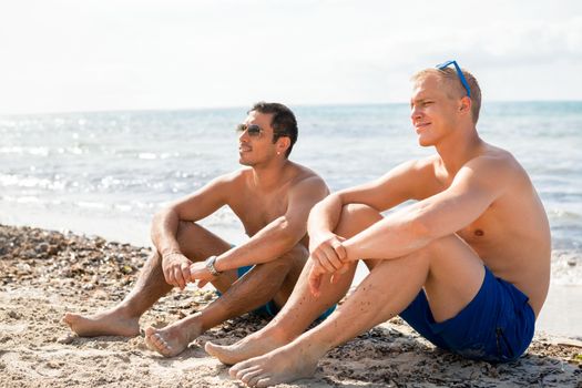 Two handsome young men chatting on a beach in their swimsuits sitting side by side on the sand with their backs to the ocean enjoying a relaxing summer day at the beach