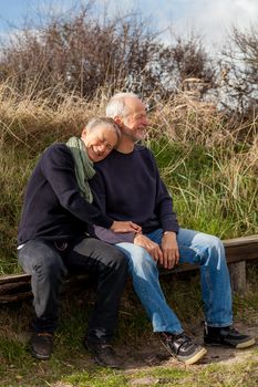 happy senior couple relaxing together in the sunshine on a wooden bench in the countryside with the one reclining full length on the seat with his head on his partners lap