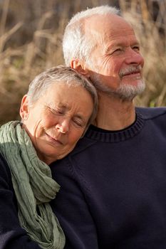 happy senior couple relaxing together in the sunshine on a wooden bench in the countryside with the one reclining full length on the seat with his head on his partners lap