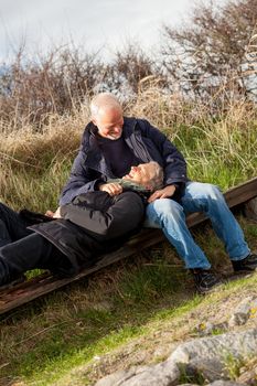 happy senior couple relaxing together in the sunshine on a wooden bench in the countryside with the one reclining full length on the seat with his head on his partners lap