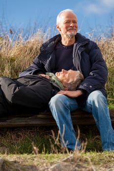 happy senior couple relaxing together in the sunshine on a wooden bench in the countryside with the one reclining full length on the seat with his head on his partners lap