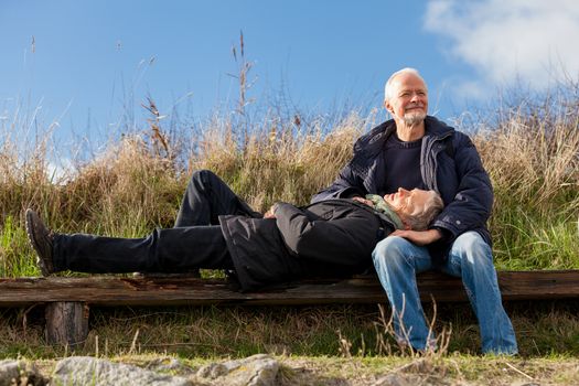 happy senior couple relaxing together in the sunshine on a wooden bench in the countryside with the one reclining full length on the seat with his head on his partners lap