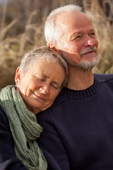 happy senior couple relaxing together in the sunshine on a wooden bench in the countryside with the one reclining full length on the seat with his head on his partners lap