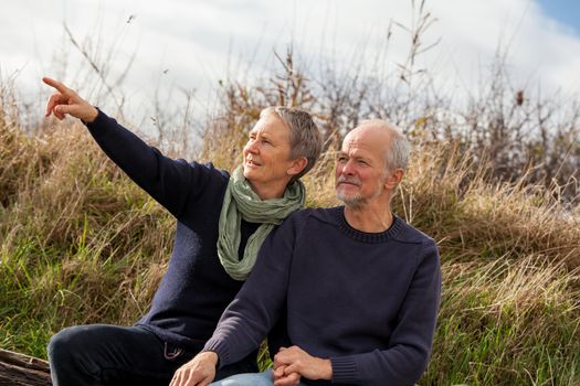 happy senior couple relaxing together in the sunshine on a wooden bench in the countryside with the one reclining full length on the seat with his head on his partners lap