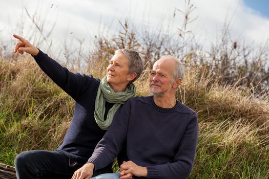 happy senior couple relaxing together in the sunshine on a wooden bench in the countryside with the one reclining full length on the seat with his head on his partners lap