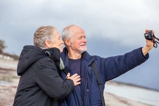 Elderly couple taking a self portrait on a compact digital camera posing in the open air and sunshine with their heads close together smiling at the lens