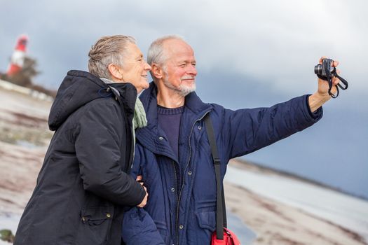 Elderly couple taking a self portrait on a compact digital camera posing in the open air and sunshine with their heads close together smiling at the lens