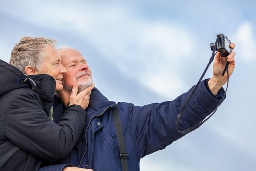 Elderly couple taking a self portrait on a compact digital camera posing in the open air and sunshine with their heads close together smiling at the lens