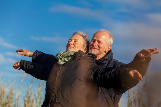 Attractive elderly couple in warm clothing standing clue together with outstretched arms, closed eyes and laughing smile against a blue sky embracing and celebrating the sun