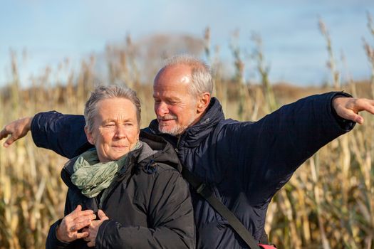 Attractive elderly couple in warm clothing standing clue together with outstretched arms, closed eyes and laughing smile against a blue sky embracing and celebrating the sun