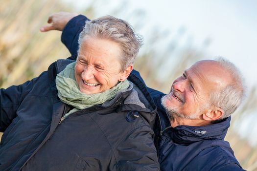 Attractive elderly couple in warm clothing standing clue together with outstretched arms, closed eyes and laughing smile against a blue sky embracing and celebrating the sun