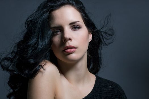 Dark moody portrait of a sultry beautiful woman with long black hair wearing a stylish off the shoulder top , head and shoulders against a grey studio background