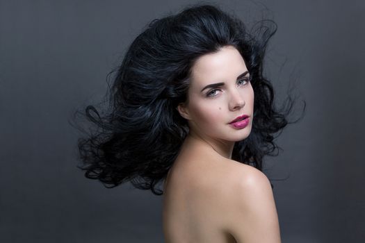 Atmospheric studio portrait of a beautiful black haired woman with a gentle serene expression gazing over her naked shoulder directly at the camera, close up head and shoulders against black