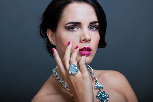 Beautiful sophisticated dark haired woman wearing elegant showy gemstone jewellery posing with bare shoulders and her hand raised gracefully to her downcast eyes with a serene expression