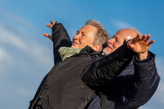 Attractive elderly couple in warm clothing standing clue together with outstretched arms, closed eyes and laughing smile against a blue sky embracing and celebrating the sun