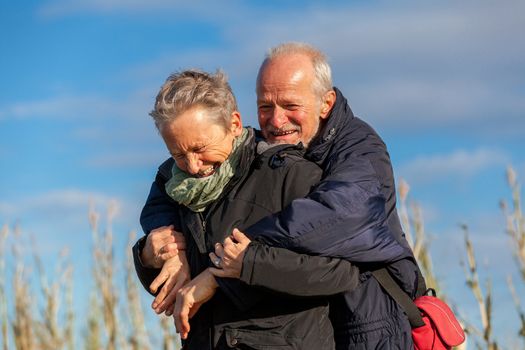 Attractive elderly couple in warm clothing standing clue together with outstretched arms, closed eyes and laughing smile against a blue sky embracing and celebrating the sun