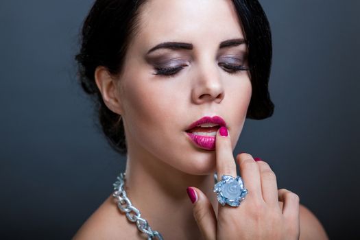 Beautiful sophisticated dark haired woman wearing elegant showy gemstone jewellery posing with bare shoulders and her hand raised gracefully to her downcast eyes with a serene expression