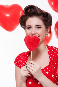 Beautiful retro woman with a French roll hairstyle celebrating Valentines Day dressed in a red polka dot dress and holding a red heart while looking at the camera with a gentle smile