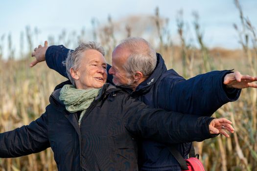 Attractive elderly couple in warm clothing standing clue together with outstretched arms, closed eyes and laughing smile against a blue sky embracing and celebrating the sun