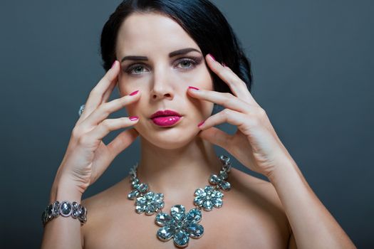 Beautiful sophisticated dark haired woman wearing elegant showy gemstone jewellery posing with bare shoulders and her hand raised gracefully to her downcast eyes with a serene expression