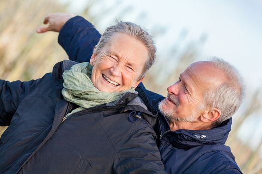 Attractive elderly couple in warm clothing standing clue together with outstretched arms, closed eyes and laughing smile against a blue sky embracing and celebrating the sun