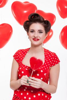 Beautiful retro woman with a French roll hairstyle celebrating Valentines Day dressed in a red polka dot dress and holding a red heart while looking at the camera with a gentle smile