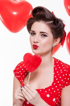 Beautiful retro woman with a French roll hairstyle celebrating Valentines Day dressed in a red polka dot dress and holding a red heart while looking at the camera with a gentle smile