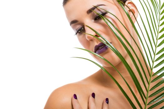 Portrait of a sensual beautiful naked woman holding a fresh palm frond with her hand with manicured purple nails resting gracefully on her shoulder as she looks down with a serious serene expression