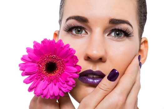 Close up facial portrait of a beautiful young brunette woman in purple make-up with her hand resting gracefull on her cheek holding a fresh magenta Gerbera daisy, isolated on white