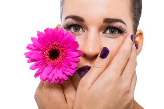 Close up facial portrait of a beautiful young brunette woman in purple make-up with her hand resting gracefull on her cheek holding a fresh magenta Gerbera daisy, isolated on white