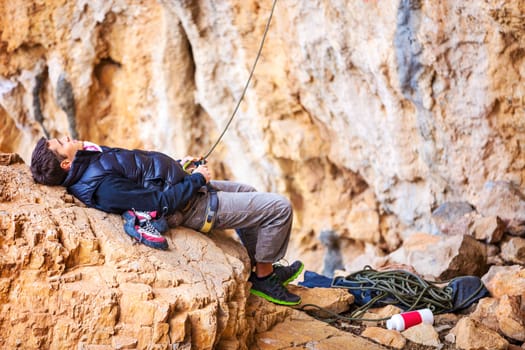 Young man lying on stone and watching leading rock climber while belaying