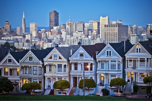 Famous Painted Ladies of San Francisco, California sit glowing amid the backdrop of a sunset and skyscrapers. 