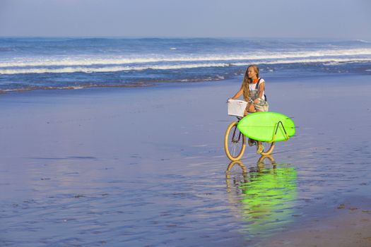 Young girl with surfboard and bicycle on the beach.