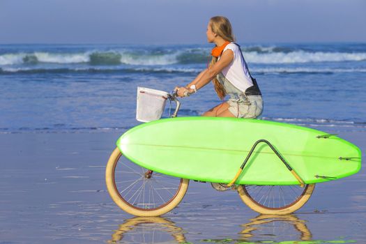 Young girl with surfboard and bicycle on the beach.