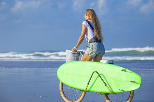 Young girl with surfboard and bicycle on the beach.