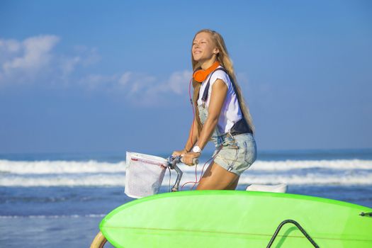 Young girl with surfboard and bicycle on the beach.