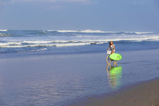 Young girl with surfboard and bicycle on the beach.