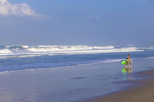 Young girl with surfboard and bicycle on the beach.