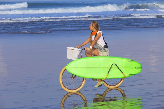 Young girl with surfboard and bicycle on the beach.