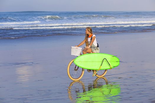 Young girl with surfboard and bicycle on the beach.