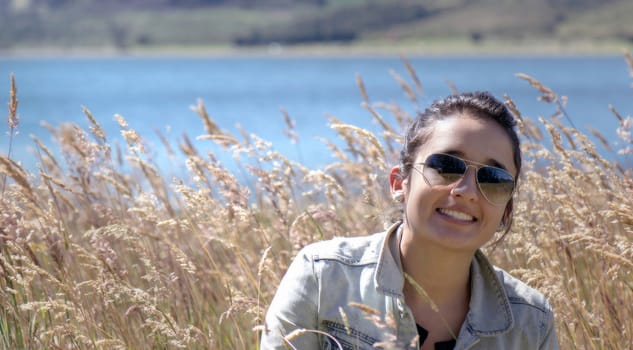 Nice young woman into a golden grass field, she showing happy with a big smile, in the background it can observe a blue lake and the slope of a hill 