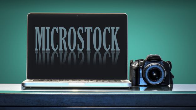 Laptop and reflex camera over a desk with aquamarine background, the computer shows a fictitious micro stock site